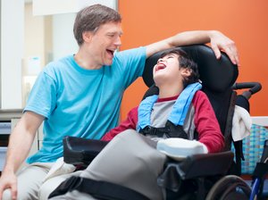 Eine Junge mit nicht genauer erkennbarer Behinderung, welcher im Rollstuhl sitzt, lacht mit einem Betreuer.