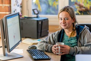 Eine Frau sitzt lächeln mit ihrer Kaffeetasse in der Hand an einen Bürotisch am Computer.