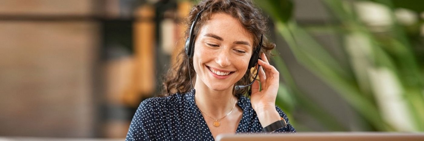 Eine Frau sitz an ihrem PC, sie trägt ein Headset telefoniert und lächelt dabei 