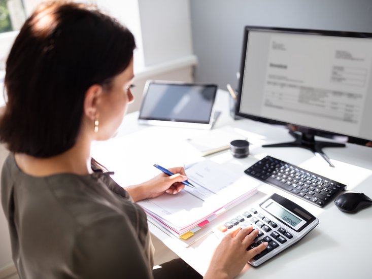 Eine Frau sitzt an in einem Büro an einen Schreibtisch an ihrem PC sie schreibt sich Notizen mit und benutzt einen Taschenrechner. 