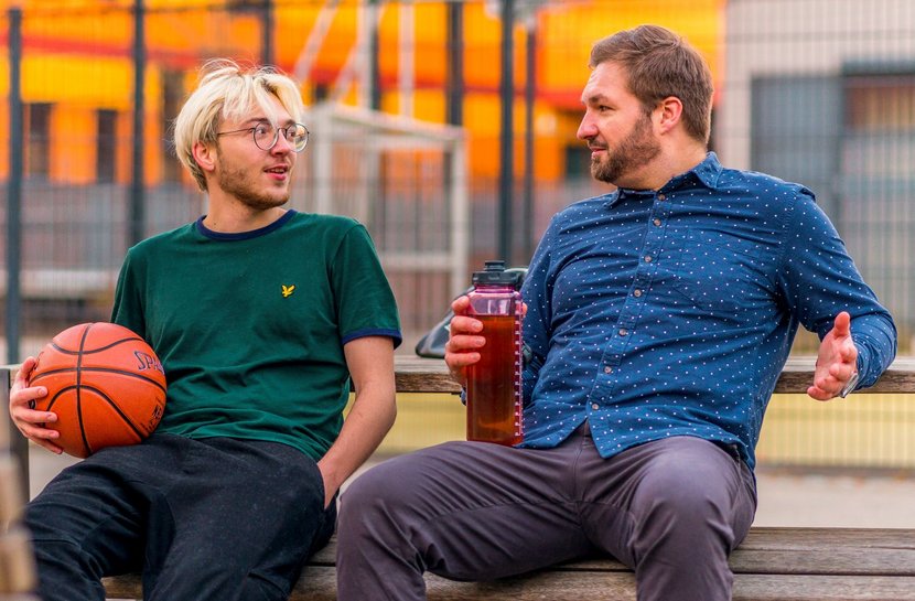 2 Männer sitzen draußen auf einer Bank. Sie sind auf einem Sportplatz. Sie unterhalten sich beide. Der linke Mann hat einen Basketball in der Hand. Der andere Mann hält seine Trinkflasche in der Hand.