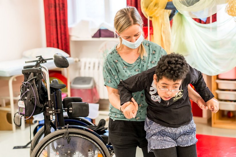 Eine Frau steht hinter einem Kind mit nicht genauer erkennbarer Behinderung. Sie hält das Kind an den Händen, sie machen zusammen eine Gehübung. Im Hintergrund steht der Rollstuhl des Kindes.